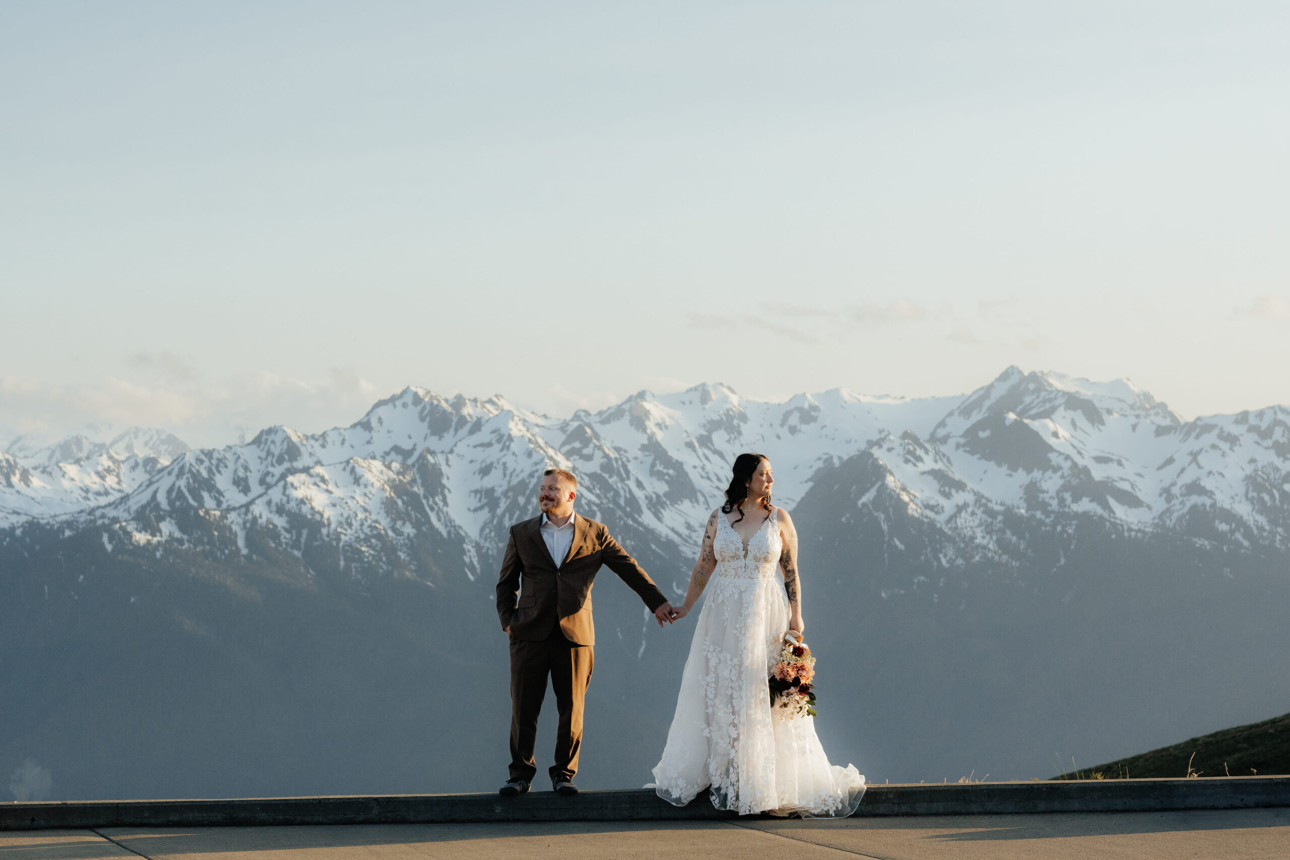 Couple holds hands while standing in front of snow-capped peaks during their Hurricane Ridge elopement.