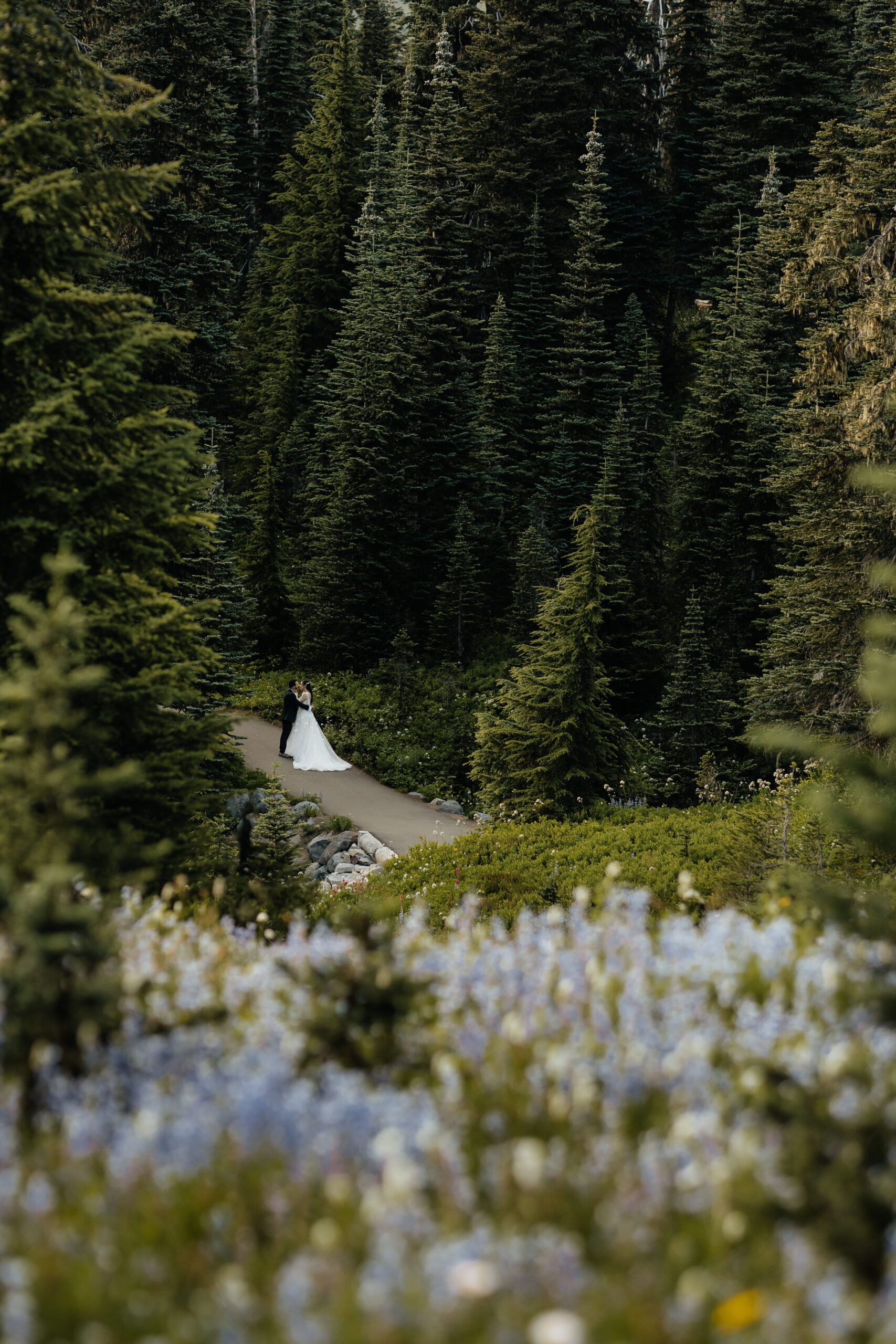 Couple stands on a trail surrounded by trees and wildflowers during their elopement at Mount Rainier, Washington.