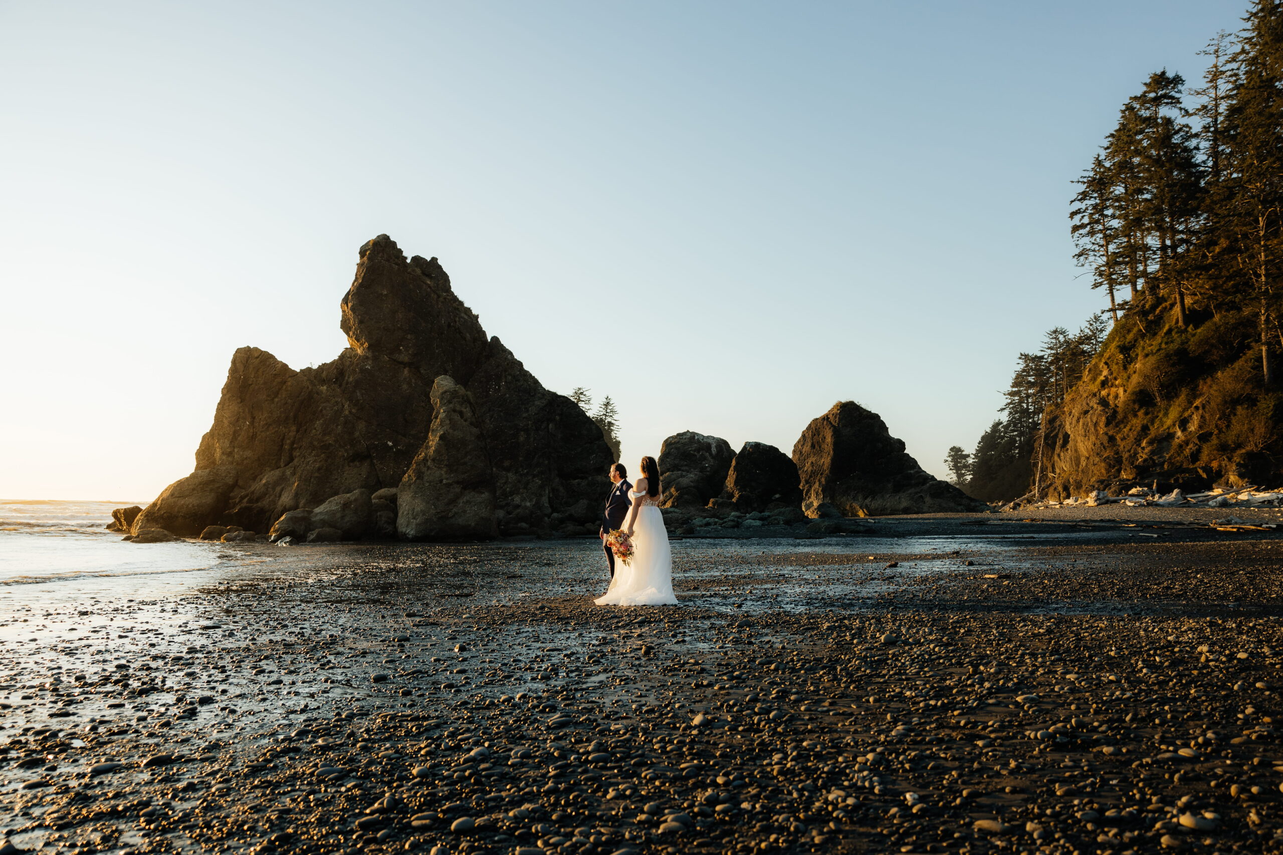 Newlyweds stand on a pebble beach next to a sea stack during their Ruby Beach elopement on the Olympic Peninsula.