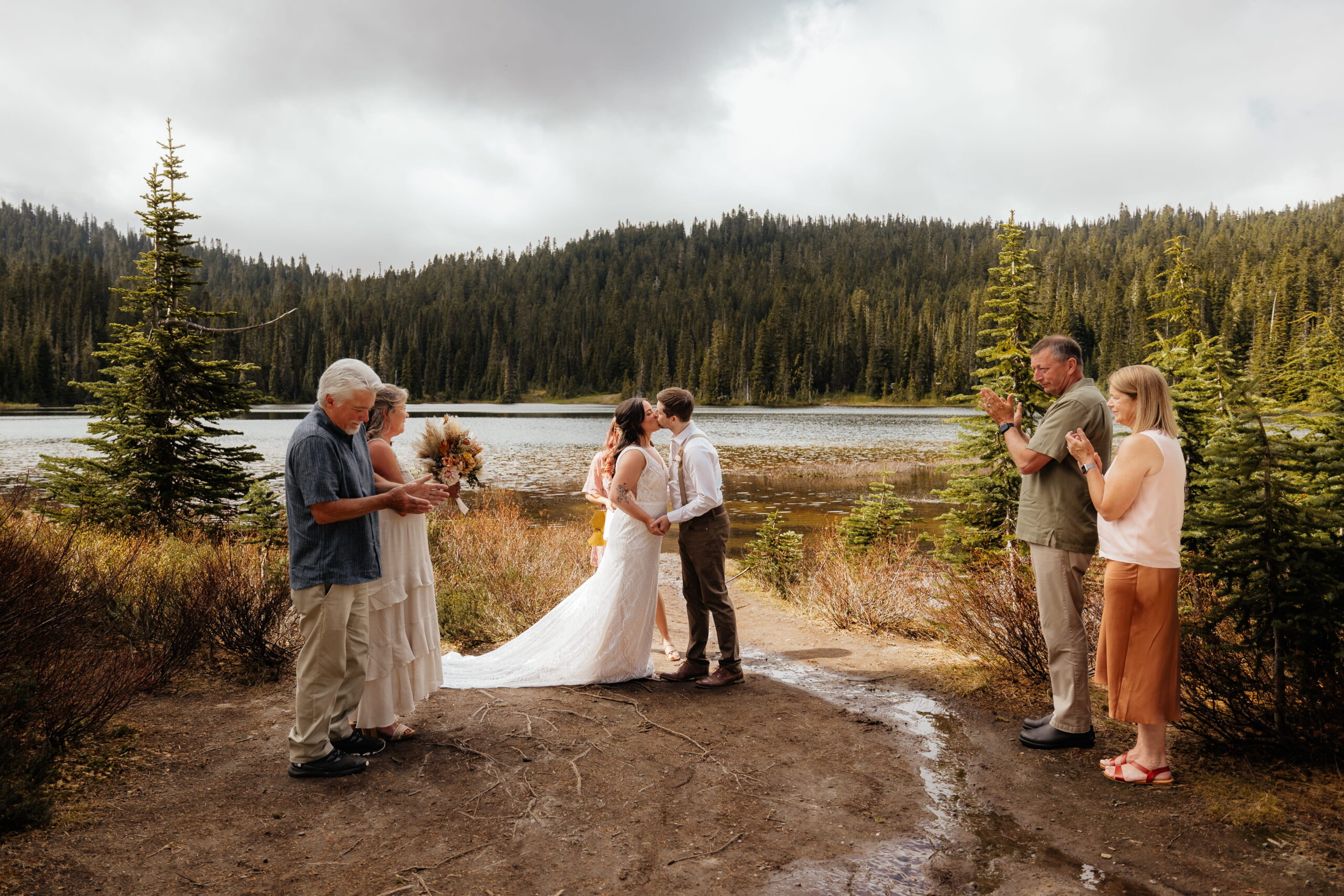 Newlyweds kiss while guests clap during their Mount Rainier wedding ceremony by a lake.