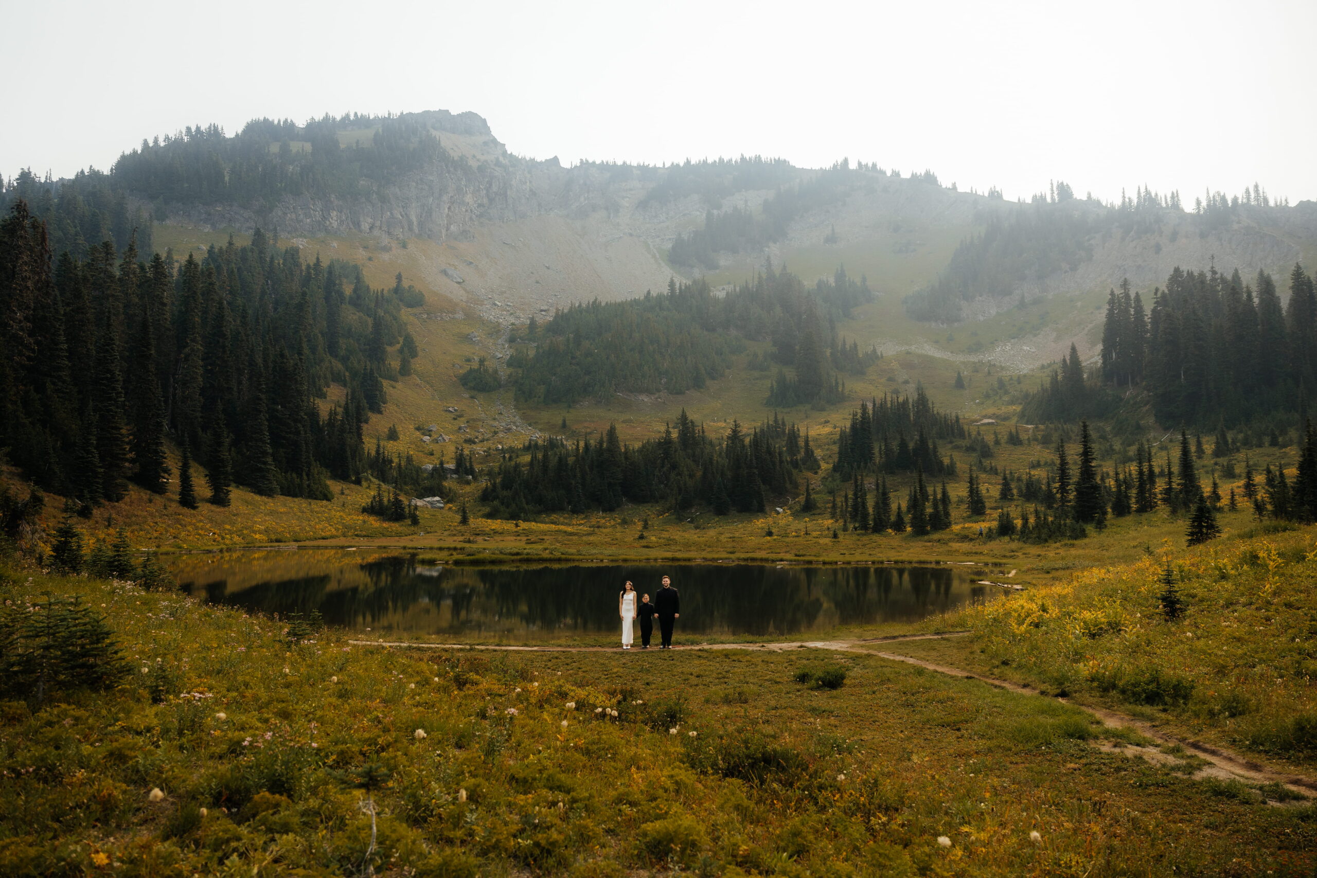 Bride and groom stand by an alpine lake with their son during their Sunrise Mt Rainier elopement.