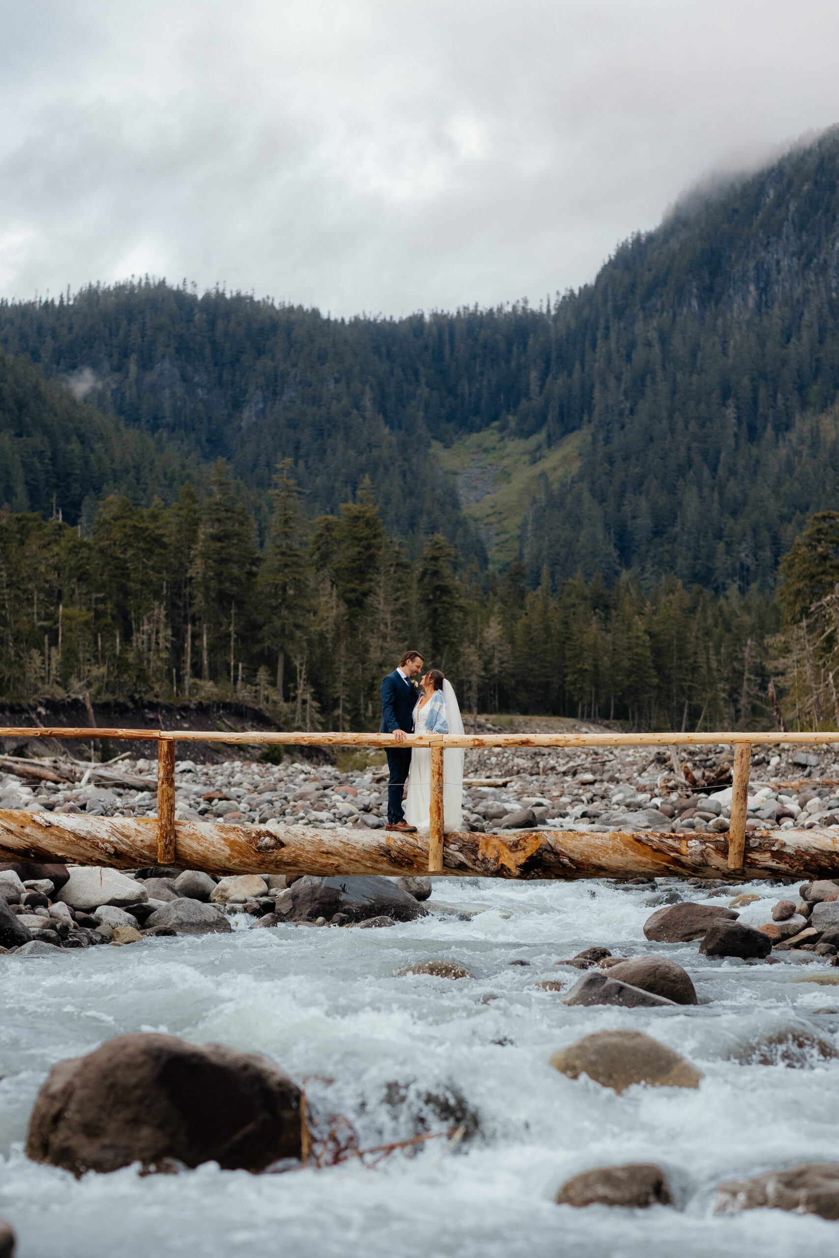 Newlyweds kiss on a log bridge over a rushing river during their spring elopement at Mt. Rainier.
