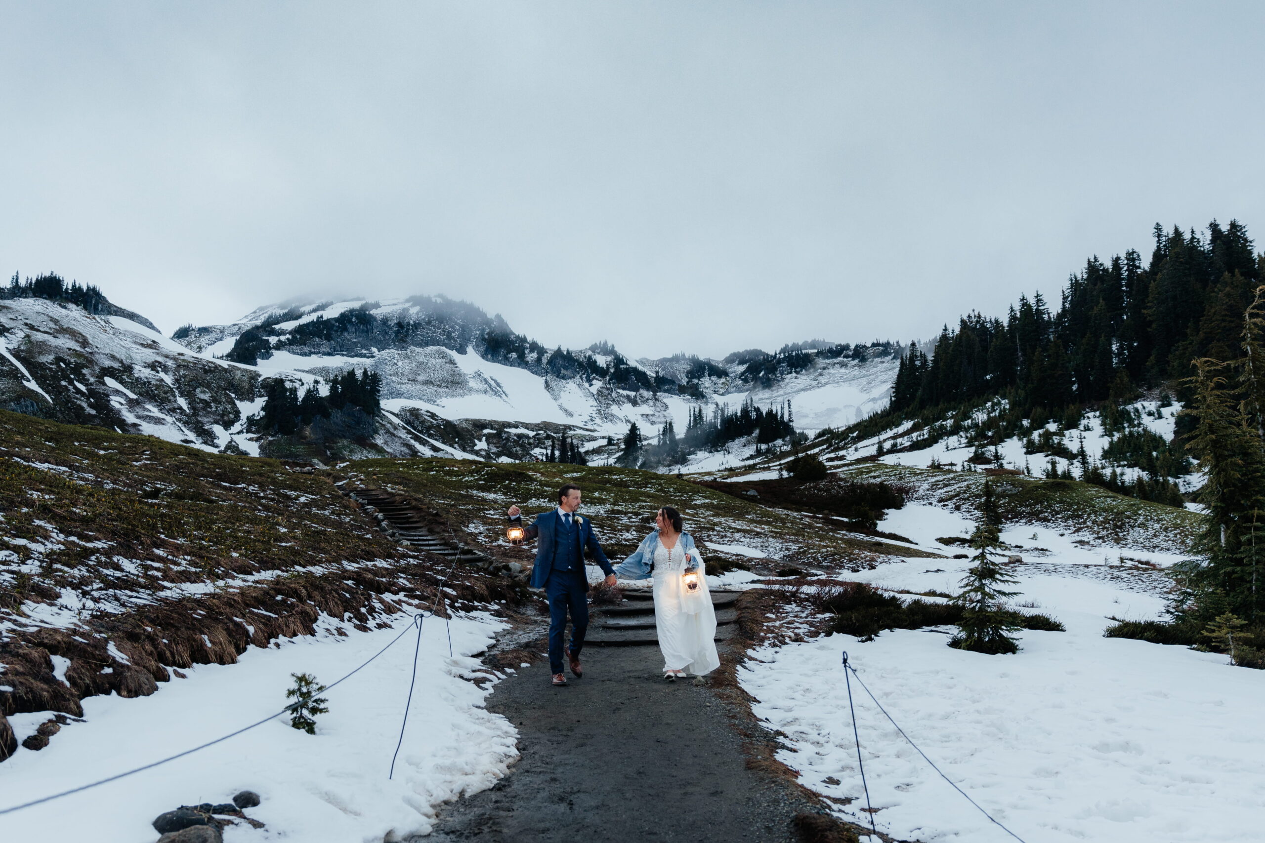 Newlyweds carry lanterns down a snowy hiking trail at Mt Rainier National Park.