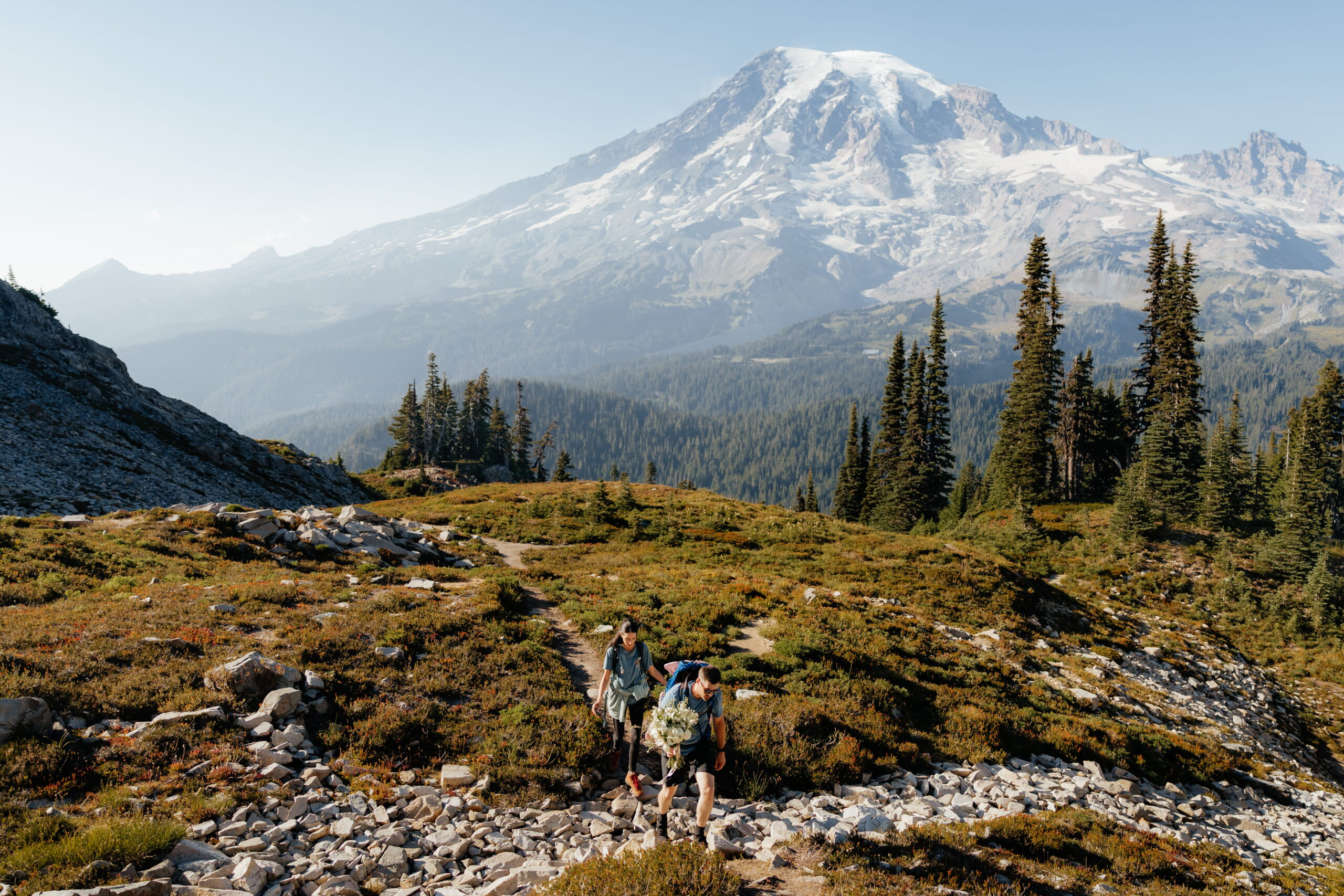 Couple hikes with wedding attire and flowers up a mountain trail at Mt Rainier National Park.